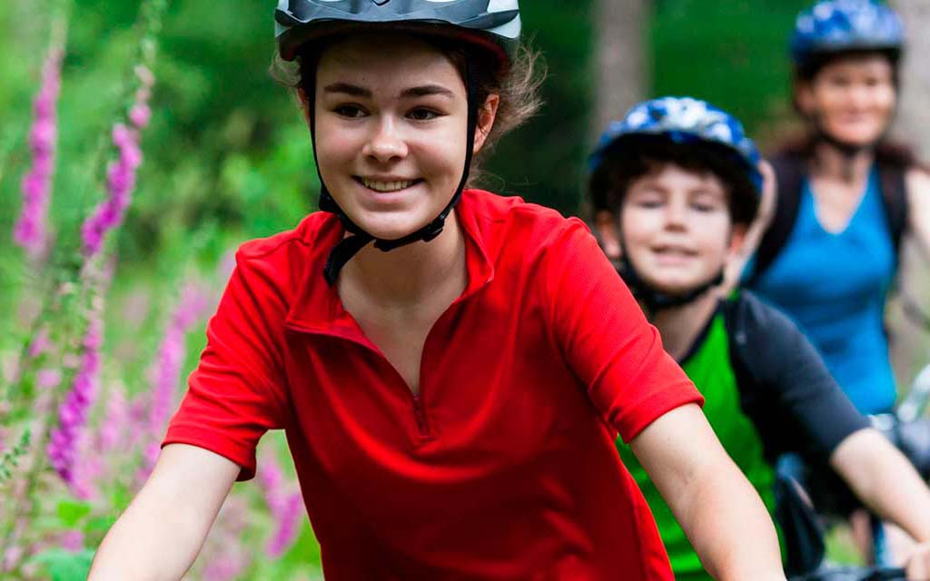 Au premier plan, une jeune fille habillé en rouge avec un casque sur la tête, fait du vélo. Au second plan, un petit garçon et sa mère en vélo également.