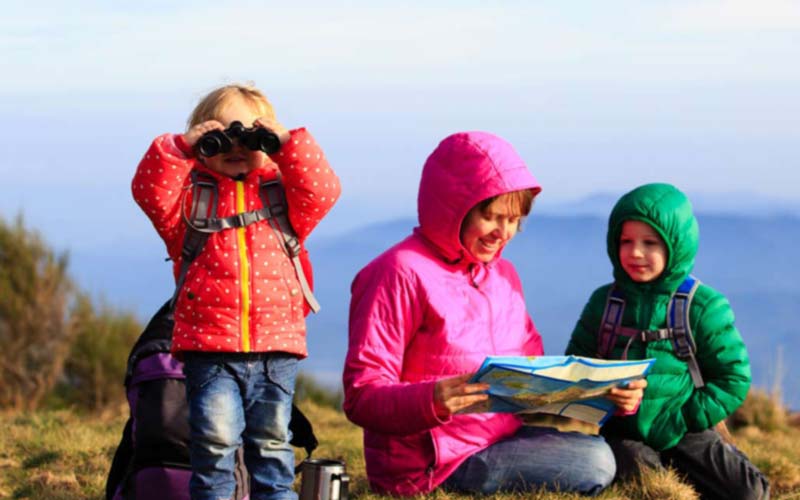 Une mère avec sa fille et son fils à la montagne, habillés avec des vêtements chauds, la petite fille regarde dans une paire de jumelles et la mère regarde une carte.