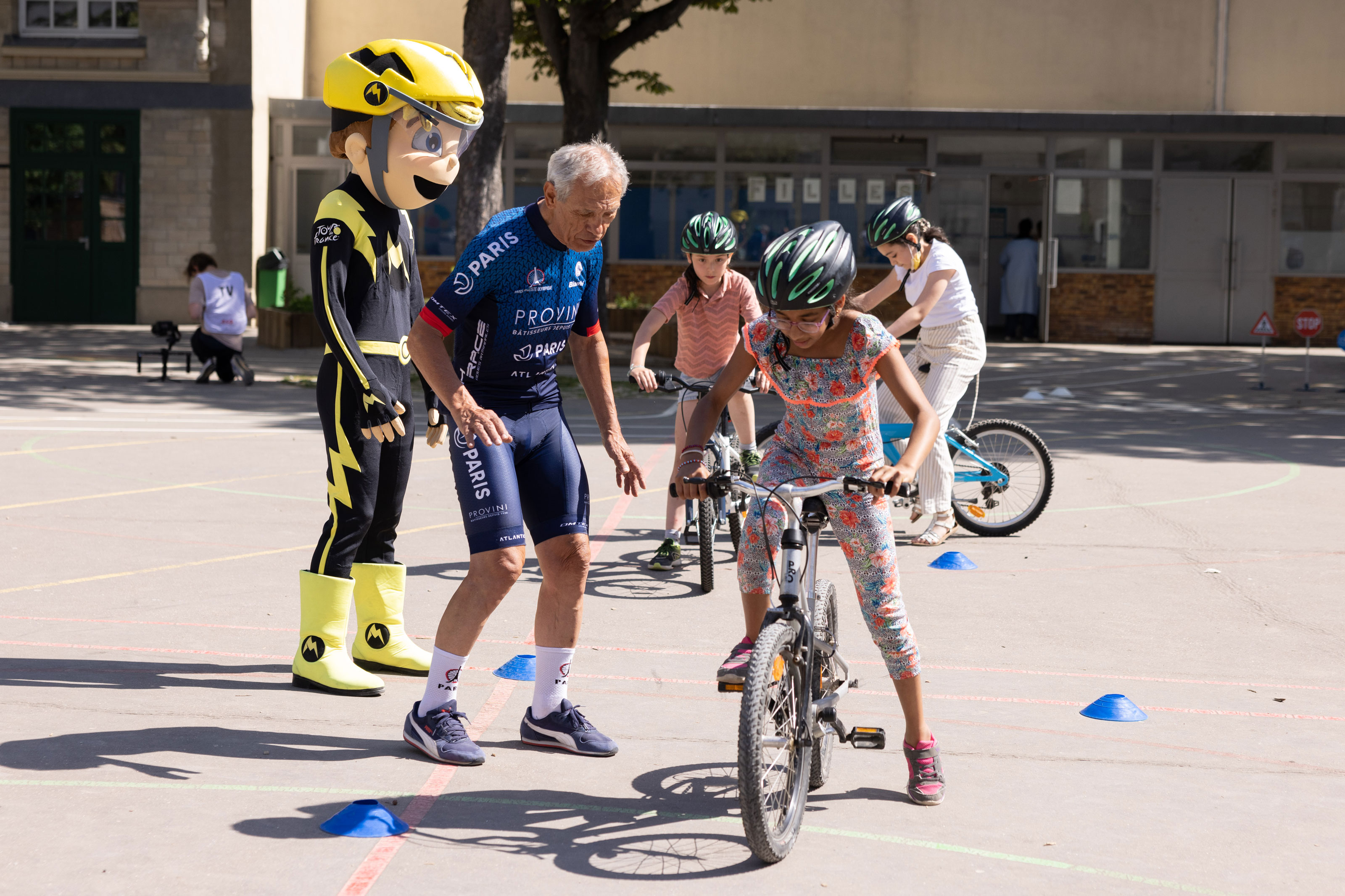 Un enseignant apprenant à des élèves à rouler à vélo dans une cour de l'école avec la mascotte du dispositif.