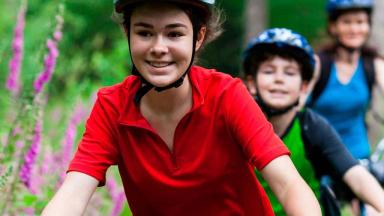 Au premier plan, une jeune fille habillé en rouge avec un casque sur la tête, fait du vélo. Au second plan, un petit garçon et sa mère en vélo également.